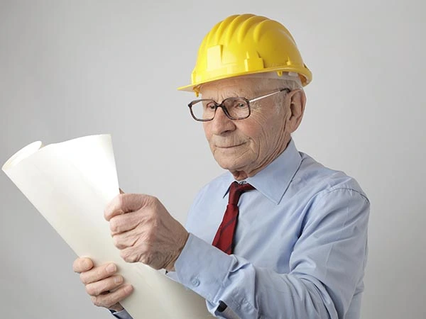 A man in business attire wearing a hard hat holding a large rolled up paper.