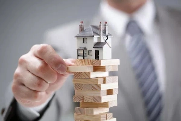 A small house set on top of a Jenga tower with a person in a business suit pulling a block from the tower.