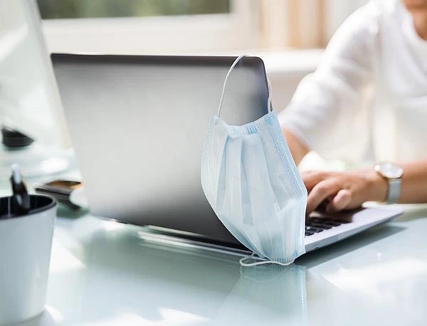 Someone working on a laptop with a face mask hanging from the top corner.